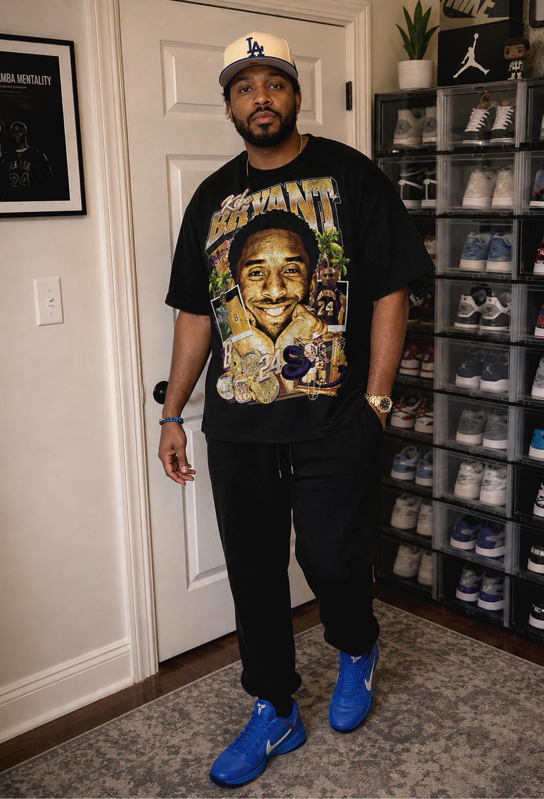 A man stands in a room wearing a black graphic t-shirt with a large portrait of Kobe Bryant, black sweatpants, blue Nike sneakers, and a Los Angeles Dodgers cap. A wall-mounted framed print is on the left, and a shoe display rack filled with sneakers is on the right.
