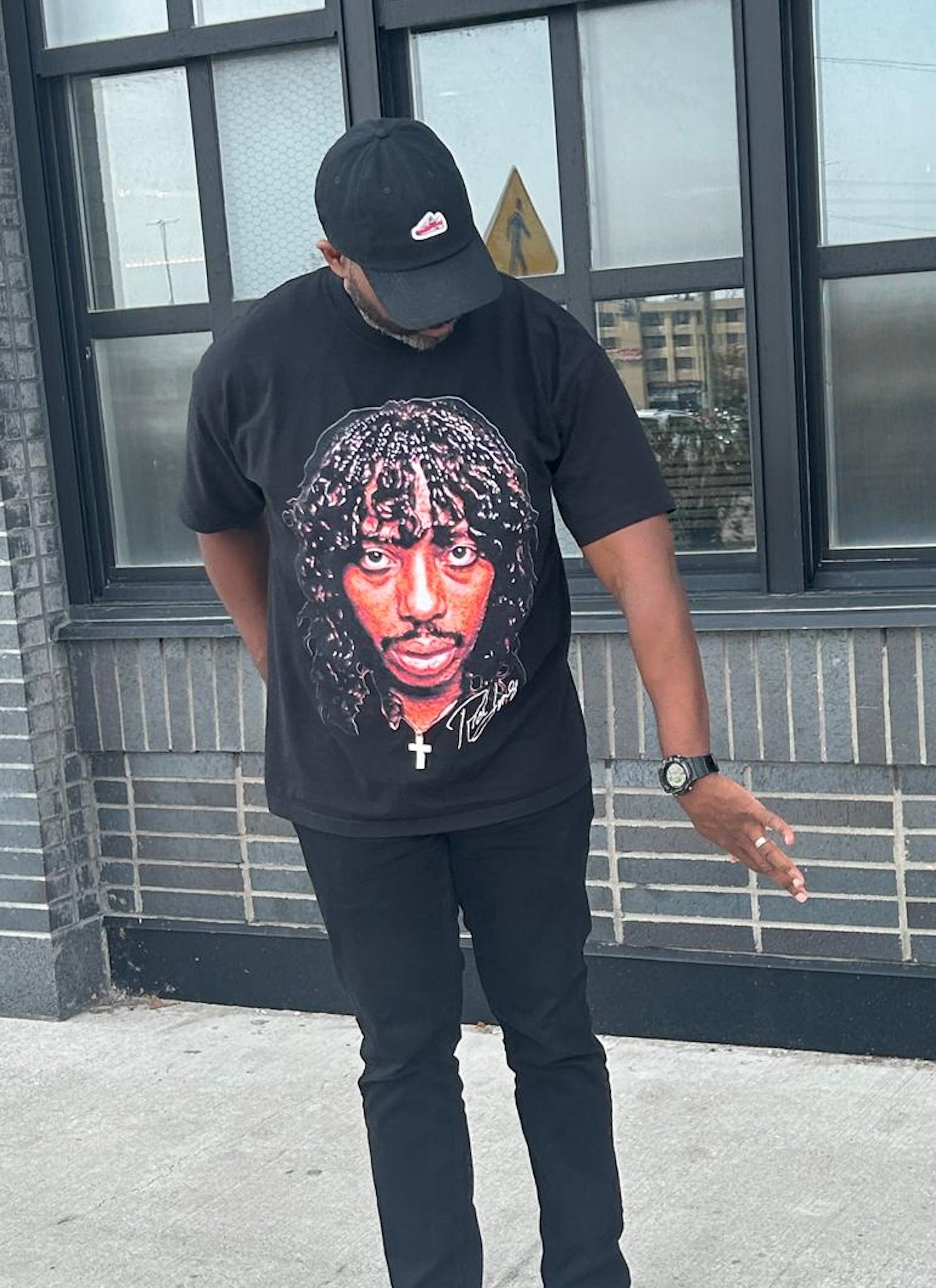 Person wearing a black t-shirt with a Rick James design, standing outside a building.