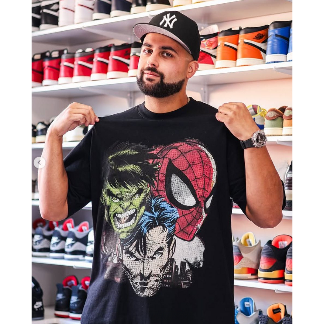 A man wearing a black graphic t-shirt featuring Marvel characters (Hulk, Spider-Man, and Daredevil) holds up the shirt to show the front print. He is wearing a black New York Yankees cap and standing in front of shelves filled with sneakers.