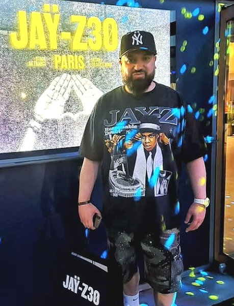 A man wearing a black graphic t-shirt with 'JAYZ' and a New York Yankees cap stands in front of a display featuring 'JAĻ-Z30 PARIS'.