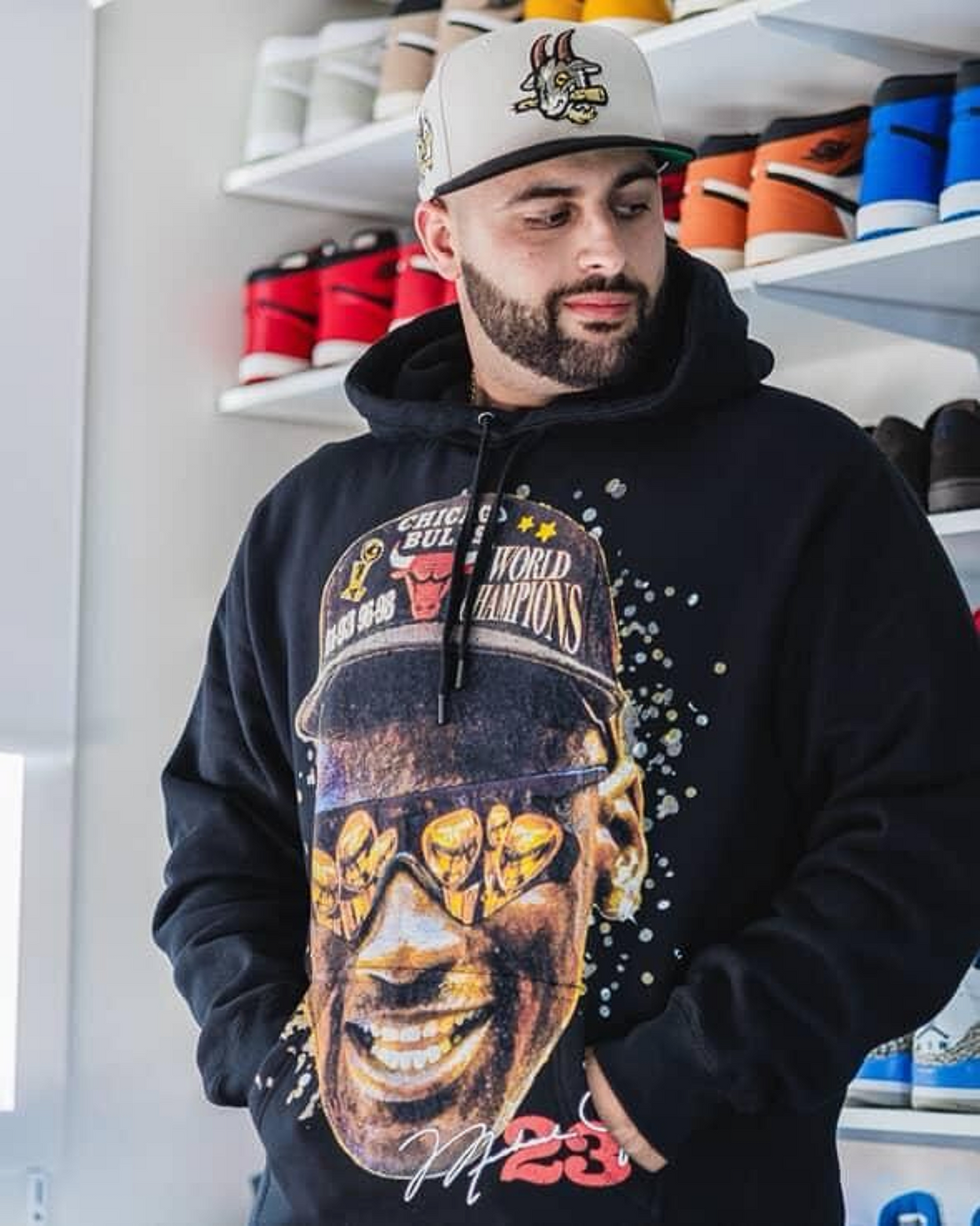A man wearing a black hoodie with a large graphic print of a basketball player wearing a Chicago Bulls championship cap, paired with a gray and black Bulls cap. The background shows shelves filled with colorful sneakers.